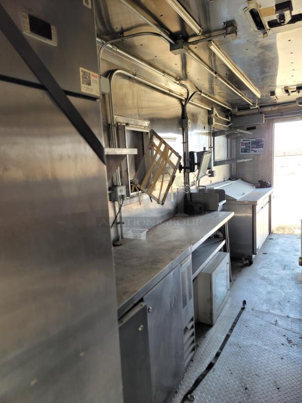 Interior of a mobile kitchen with stainless steel counters, appliances, and overhead shelves. Some wear, but clean. Suitable for food prep.