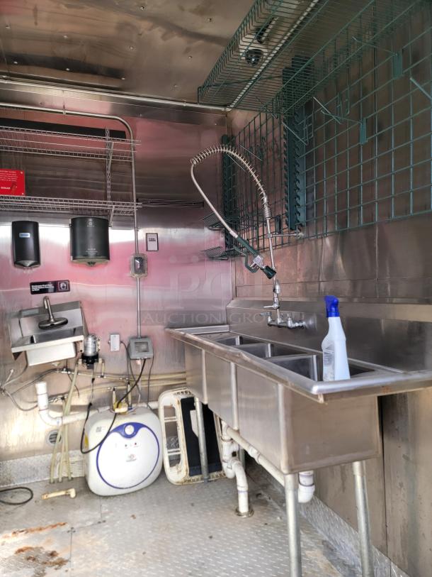 Stainless steel commercial kitchen sink with three basins, pre-rinse faucet, and storage shelves in a metal-paneled interior.