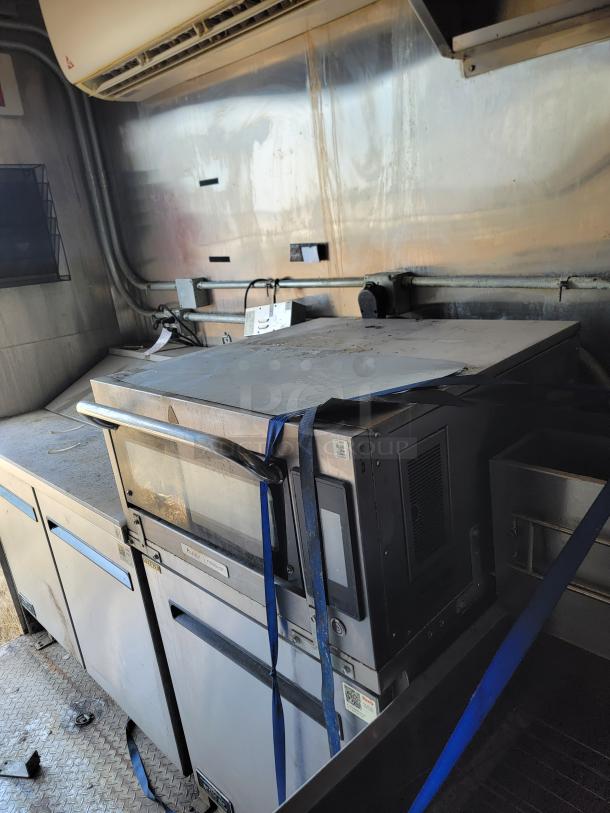 Commercial stainless steel oven and microwave combo in used condition, secured with blue straps, visible control panel.