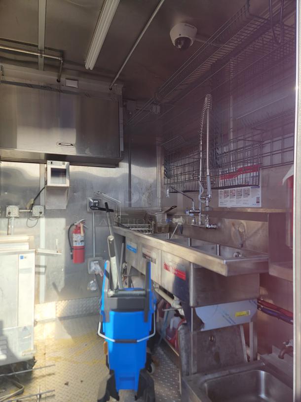 Commercial kitchen interior with stainless steel sink, industrial faucet, storage racks, mop bucket, and wall-mounted fire extinguisher.