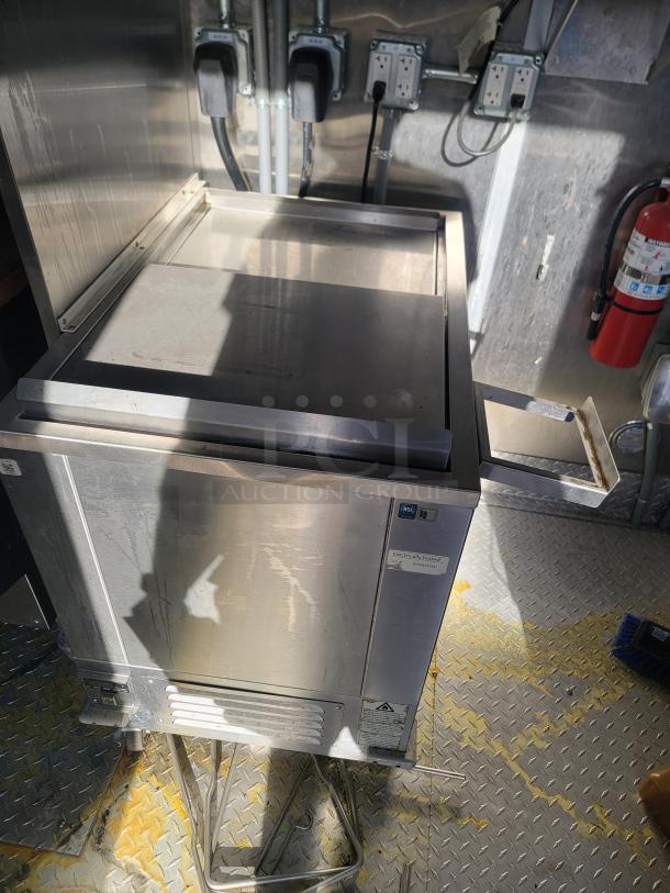 Stainless steel commercial refrigeration unit with top lid, electronic testing label, and power outlets in a utility setup.