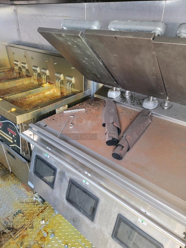 Commercial kitchen fryer with four baskets, grease residue visible. Stainless steel cover panel with folding doors. Used condition.