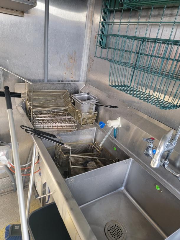 Commercial kitchen sink with fryer baskets and utensils, stainless steel, used condition, visible shelving and plumbing.
