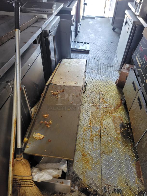 Commercial kitchen interior with stainless steel counters, diamond plate floor, broom, and visible grease spills. Used condition.