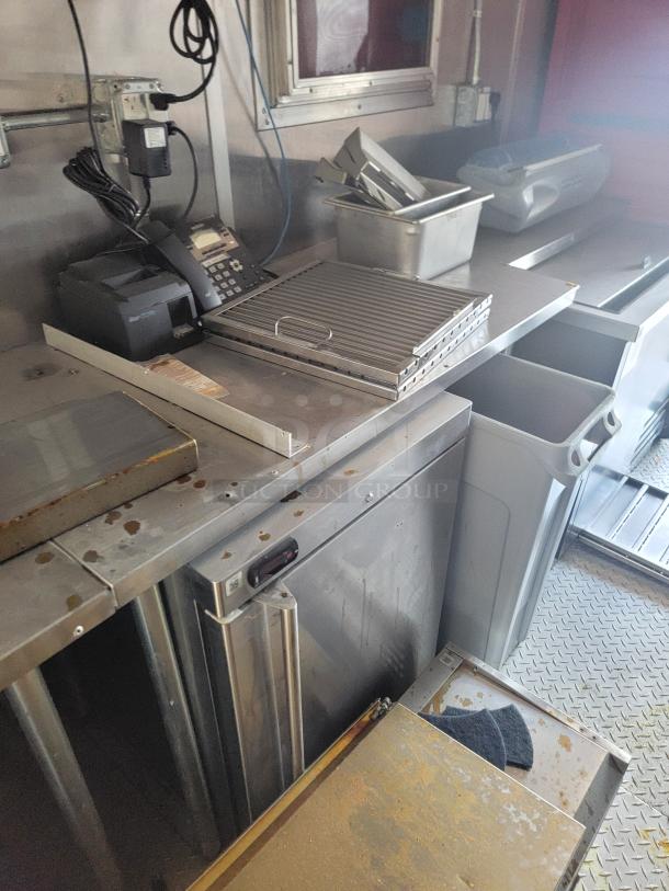 Stainless steel kitchen workstation with phone, receipt printer, metal trays, and storage bins, showing signs of use.