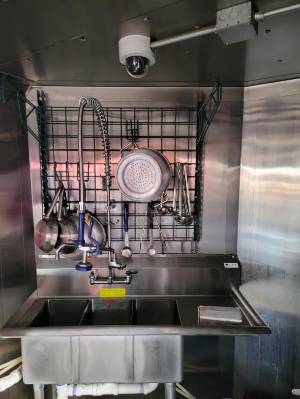 Stainless steel commercial kitchen triple sink with faucet, pot rack, and utensils. Wall-mounted security camera above. Good condition.