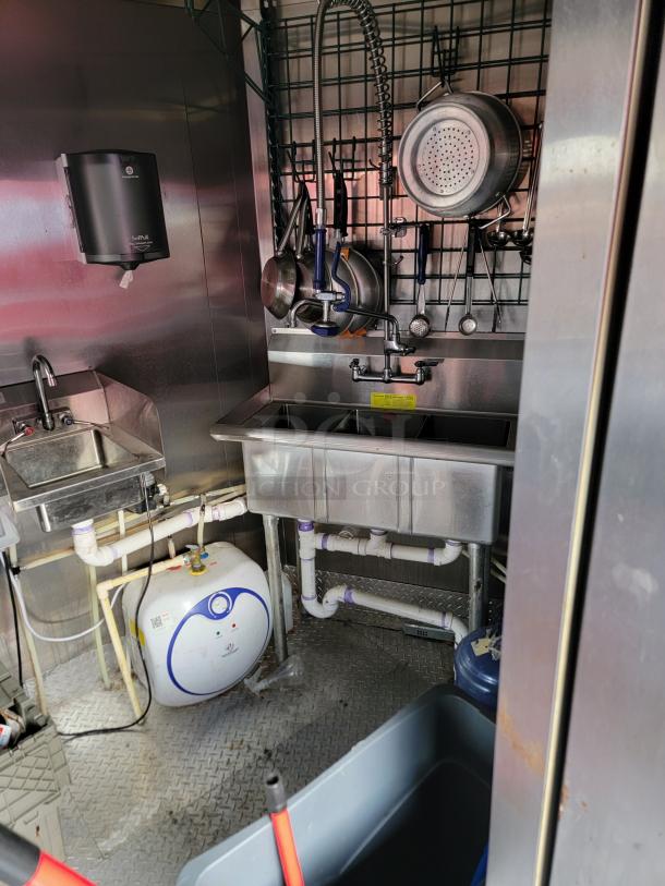Industrial kitchen sink area with stainless steel double basin, wall-mounted dish rack, and plumbing pipes. Includes sanitizer dispenser and water heater.