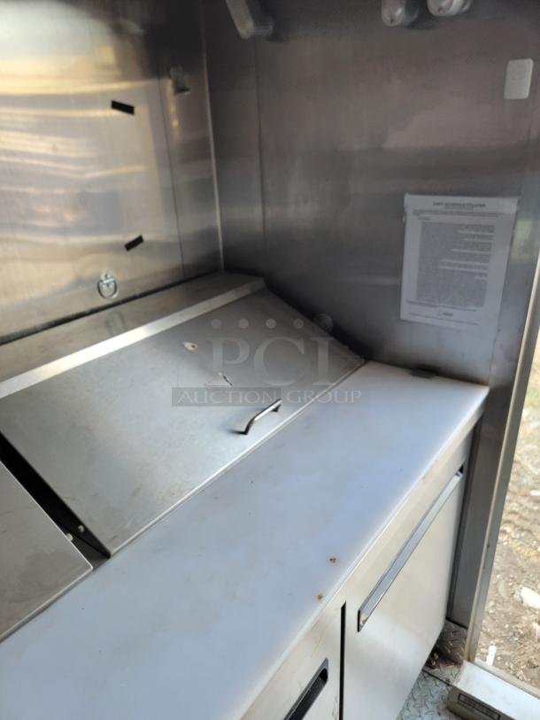 Industrial stainless steel prep station with storage, used condition, featuring a sliding lid and a document posted on the wall.