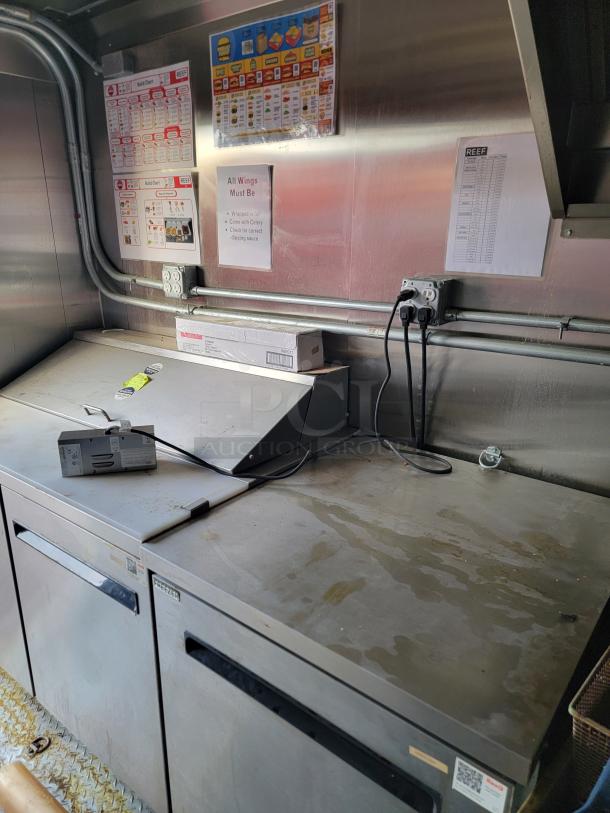 Commercial kitchen prep area with stainless steel countertops, fridge units, menus, and safety notices. Signs indicate food prep instructions.