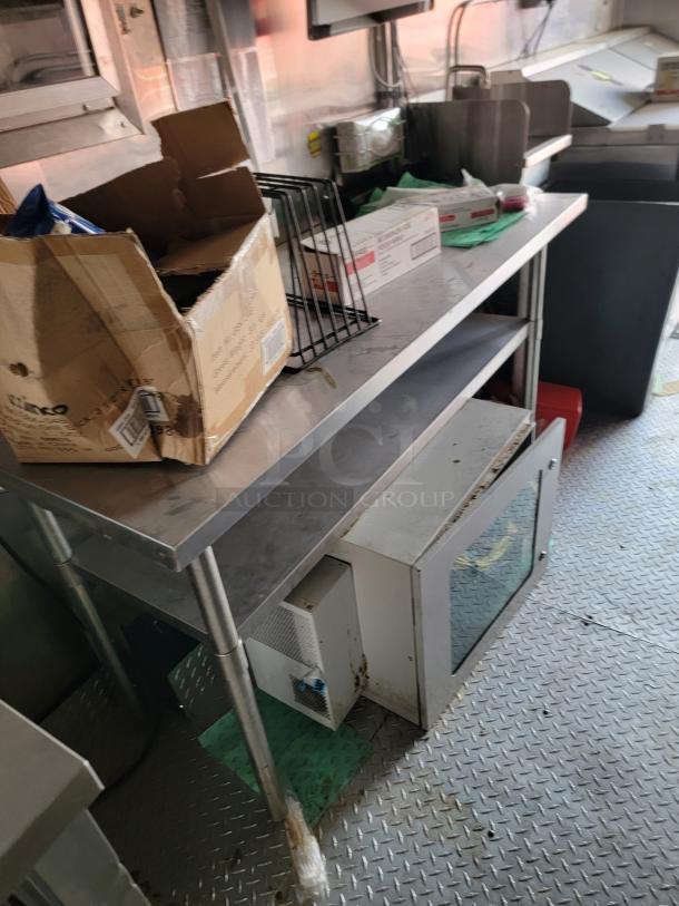 Stainless steel kitchen prep table with shelf, holds open boxes and equipment. Table shows signs of wear, suitable for commercial use.