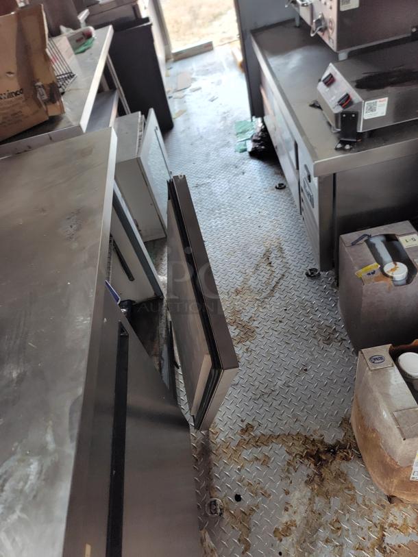 Commercial kitchen interior with stainless steel countertops, storage units, and equipment. Floor shows signs of wear.