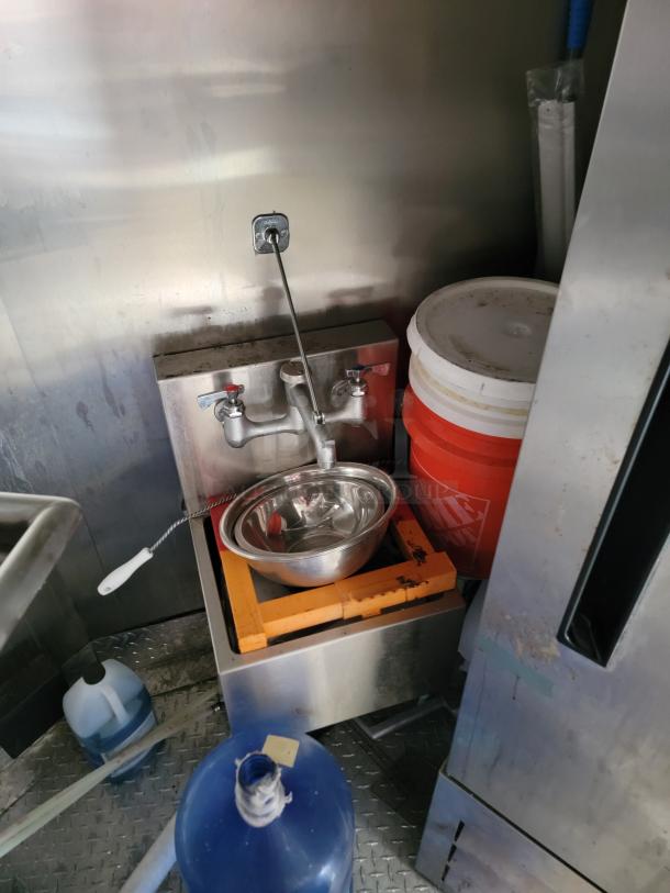 Stainless steel hand sink with double handles, mounted on a wall. Includes a metal bowl, orange crate, and visible wear.