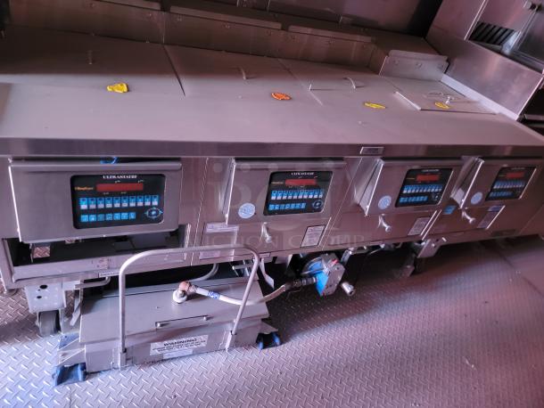Industrial kitchen fryer system with four Ultrastat25 control panels. Features stainless steel finish, digital displays, and labeled valves. Model information visible; designed for high-volume use.