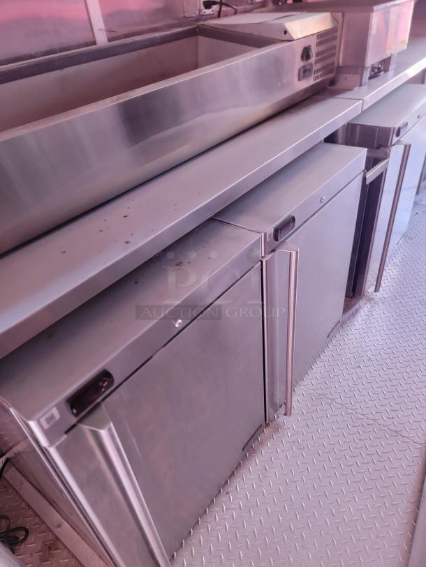 Commercial stainless steel refrigerator unit with dual doors, metal worktop, minor surface wear, ideal for kitchen use.