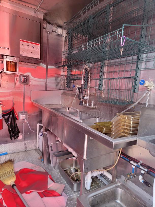 Commercial kitchen setup with stainless steel triple sink, wire shelves, and cleaning supplies. Pristine condition, ready for use.