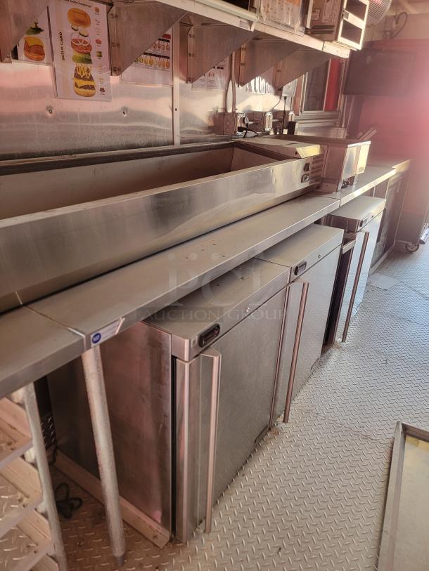 Stainless steel commercial kitchen prep station with dual refrigeration units, shelving, and condiment storage, in clean condition.