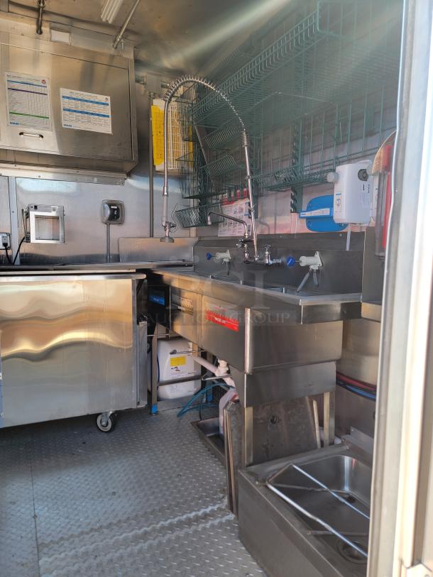 Stainless steel commercial kitchen setup with dual sinks, faucet, storage shelves, and water heater in clean condition. Ideal for food truck use.