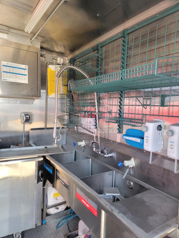 Stainless steel triple sink in a catering truck, featuring high-arc faucet, overhead shelving, and mounted soap dispensers.
