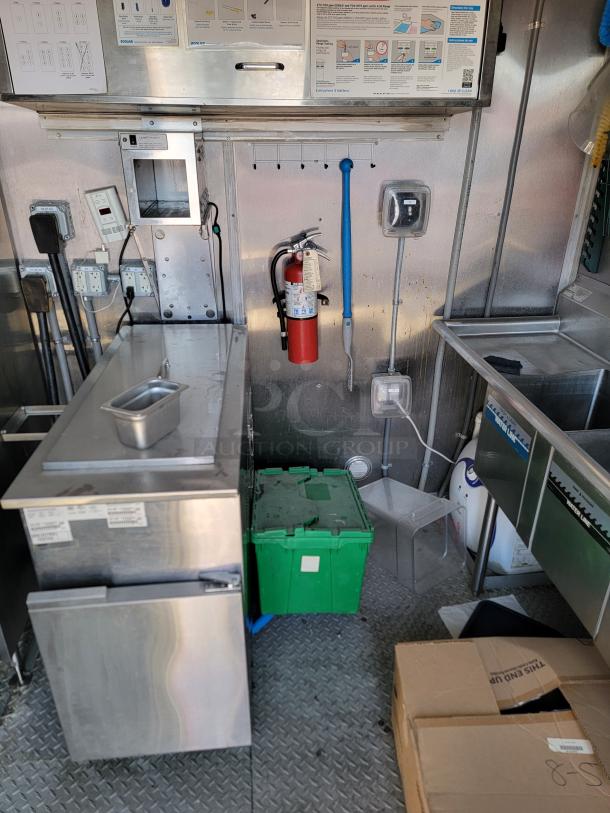Mobile kitchen interior featuring stainless steel equipment, fire extinguisher, storage, and cleaning supplies. Functional and used condition.