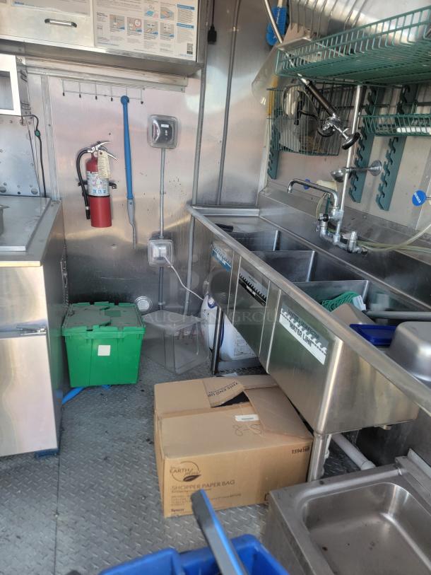 Commercial kitchen interior with stainless steel appliances, triple sink, fire extinguisher, and storage boxes. Clean condition.