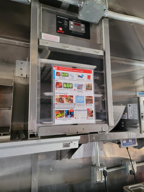 Wendy's bun holding cabinet with control panel, displaying food safety standards poster. Stainless steel finish, industrial setting.
