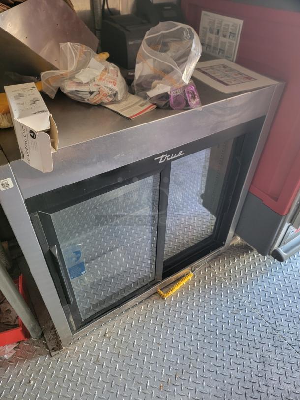 True double-door glass refrigerator, stainless steel, featuring brand logo. Shows surface wear, stored items on top.