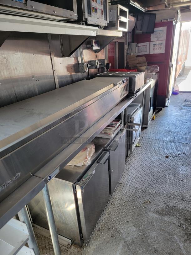 Commercial stainless steel refrigerated prep table in food truck. Features multiple storage units and seamless steel surface, in good condition.