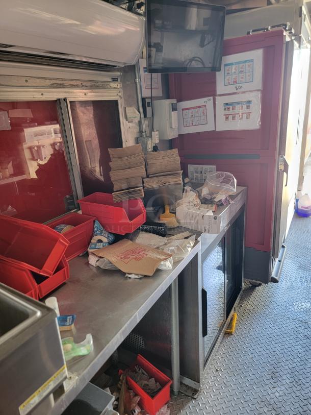 Interior of a food truck with stainless steel counters, red storage bins, paper bags, air conditioner unit, and display screen.