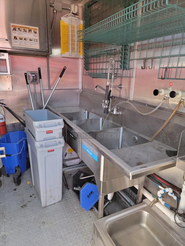 Stainless steel commercial sink with dual basins, industrial faucet, overhead green storage racks, and cleaning equipment in a utility setting.