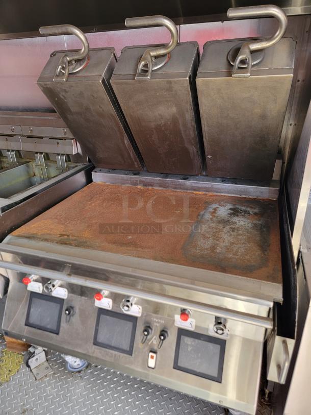 Commercial griddle with three clamshell presses, visible wear on surface, control panel with switches and screens.
