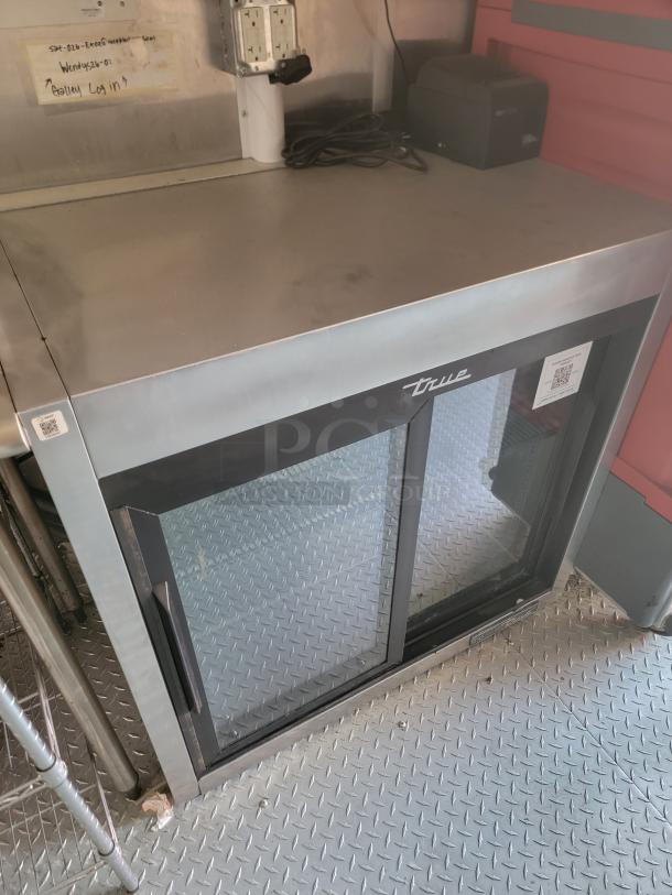 True commercial refrigerator with glass sliding doors and stainless steel top. Visible signs of use. Markings include a QR code.