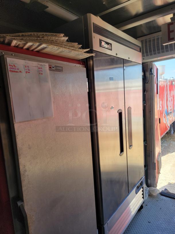 Commercial refrigerator with Migali logo in stainless steel finish. Dual-door design, includes adjacent whiteboard with notes.