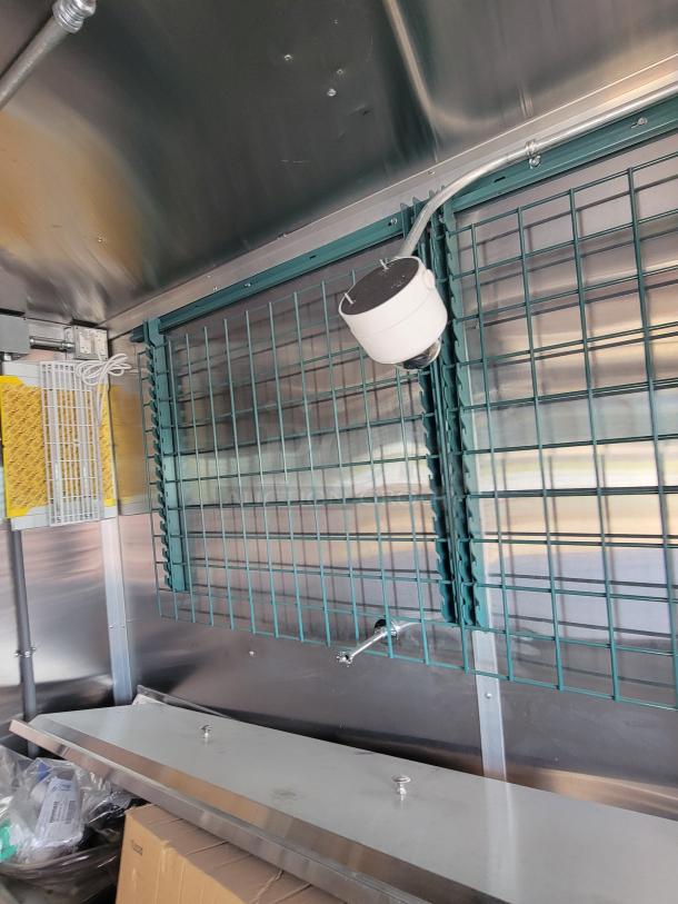 Interior view of a vehicle with metal grating on walls, a mounted white security camera, and utility shelving.