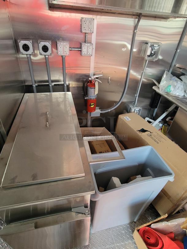 Commercial kitchen setup with stainless steel surfaces, electrical outlets, and a fire extinguisher. Includes trash bin and red mat.