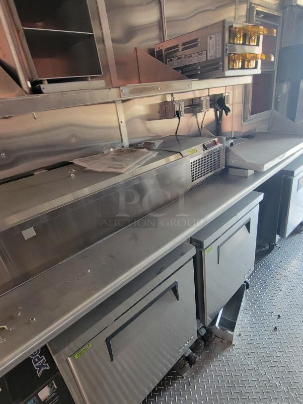 Commercial stainless steel kitchen setup with refrigeration units, wall shelving, and mounted condiments containers. Clean condition.