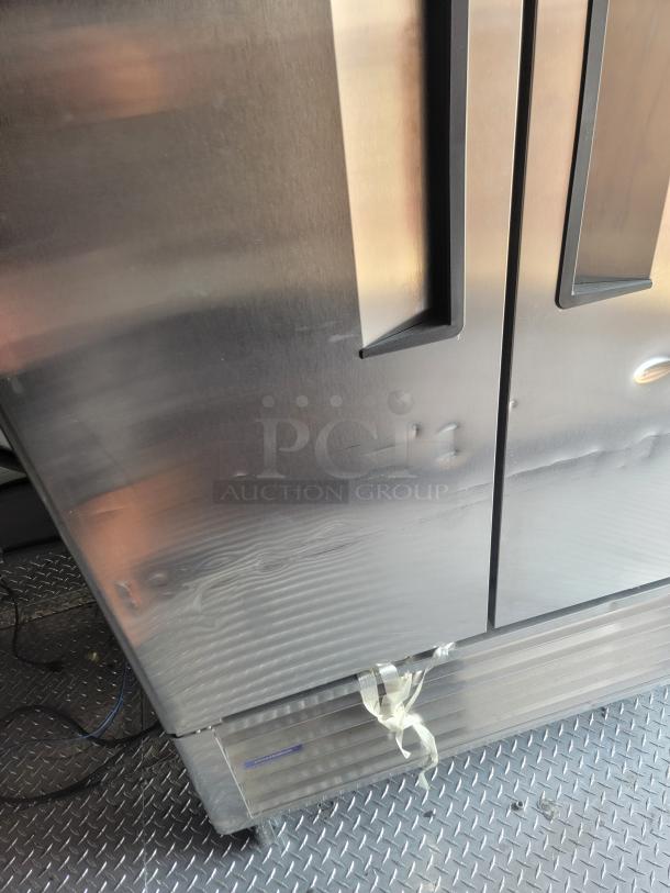 Stainless steel double-door refrigerator with visible dents. Bottom section secured with tape. Textured metal floor.