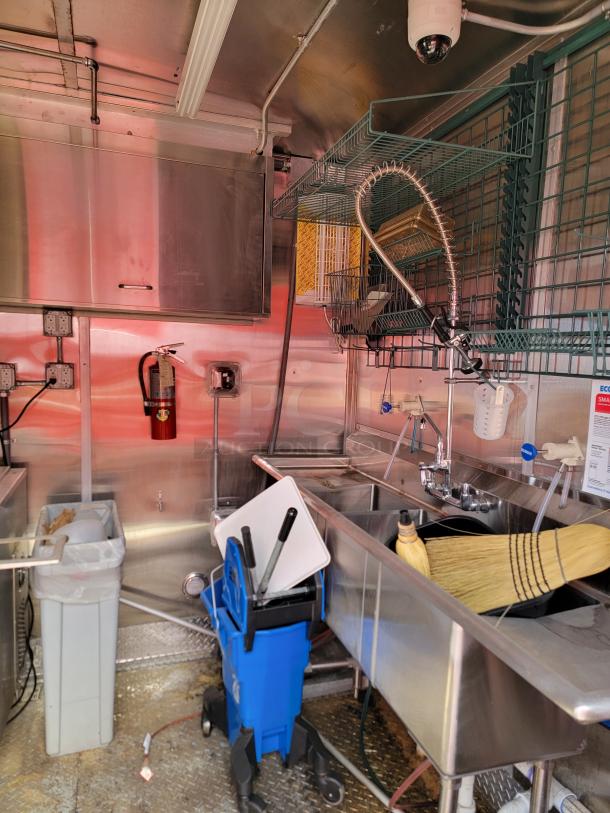 Commercial kitchen setup with stainless steel sink, faucet, and wire shelving. Includes mop bucket, broom, and trash bin.