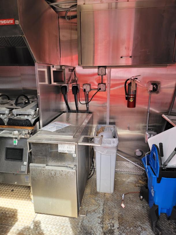 Commercial kitchen interior with stainless steel equipment, including a fire extinguisher and waste bins. Visible electrical outlets and conduits.