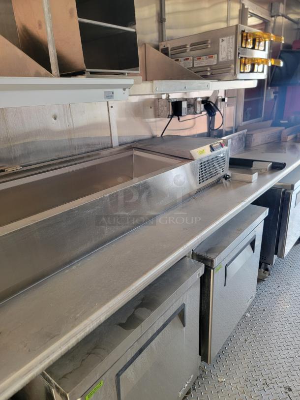 Commercial kitchen equipment setup with stainless steel countertop, built-in refrigeration units, and overhead shelving.