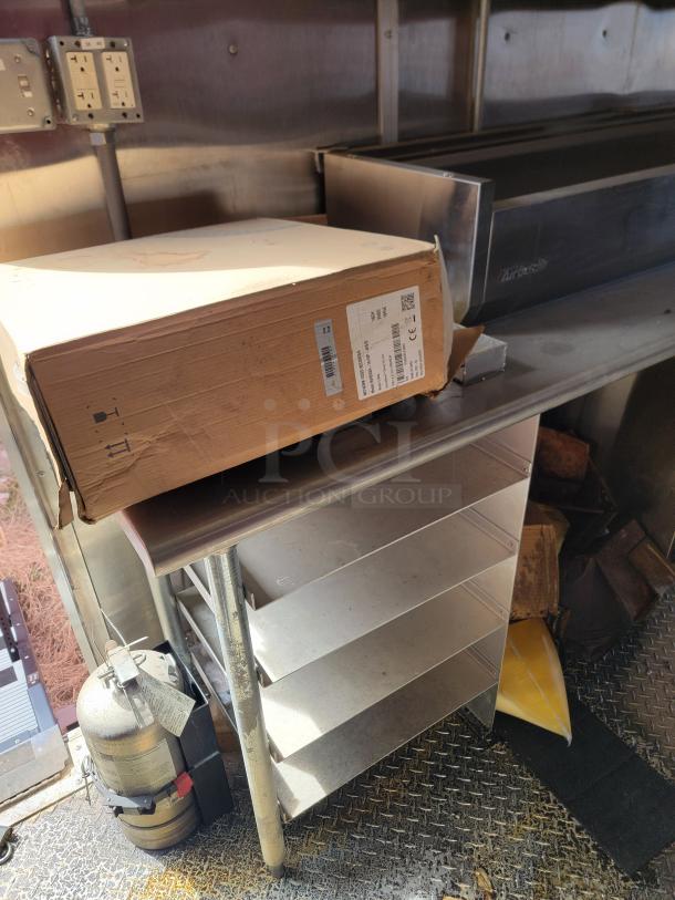 Cardboard box with product label on stainless steel table, surrounded by shelves and a fire extinguisher. Box shows minor wear.