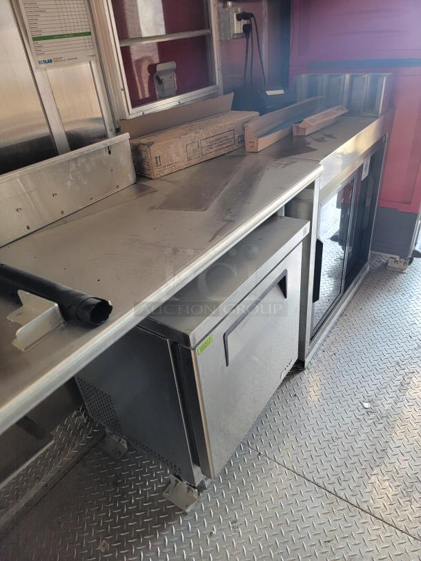 Stainless steel counter with attached shelves, showing boxes and equipment, on a textured metal floor in used condition.
