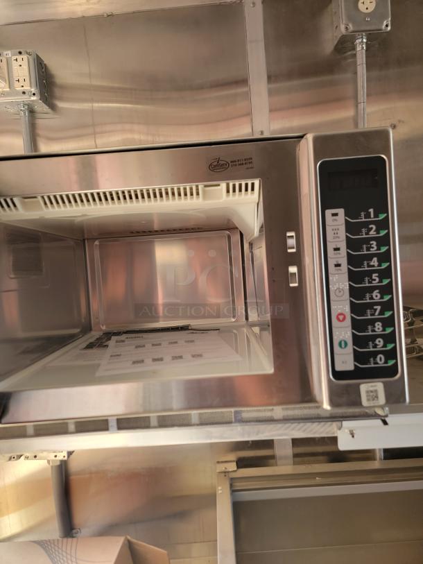 Commercial microwave, Amana model, stainless steel, digital controls, interior light. Tag shows ComServ contact info.