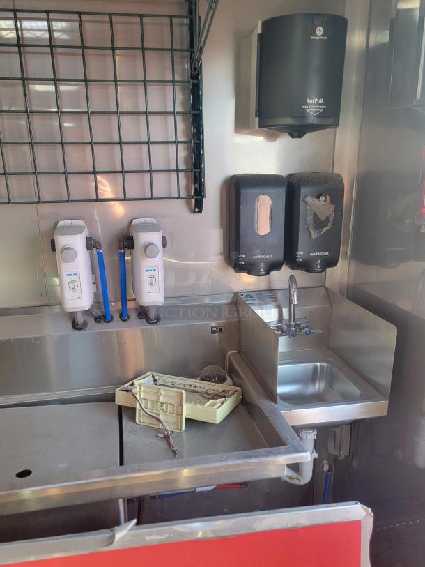 Stainless steel commercial sink with soap dispensers and a paper towel holder, featuring worn condition.