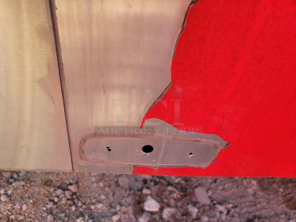 Metal surface with missing VIN, painted red and tan, shows bolt holes and exposed edges, weathered condition.