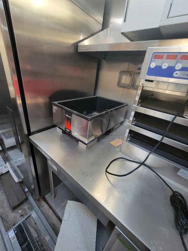 Commercial deep fryer on a stainless steel kitchen counter. Features a black frying basket, visible control panel, and power cord. Tags indicate manufacturer details.