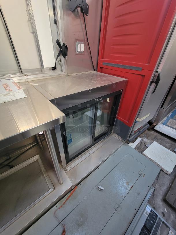 Commercial beverage cooler by True, featuring double glass doors, located in a stainless steel kitchen setup. Appears in used condition.