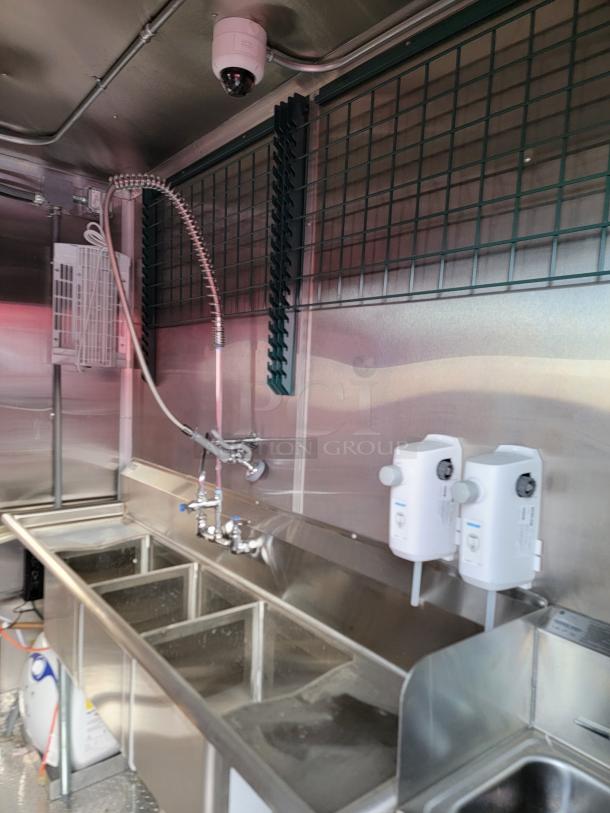 Stainless steel commercial kitchen sink with three compartments, overhead spray nozzle, and mounted soap dispensers. New condition.