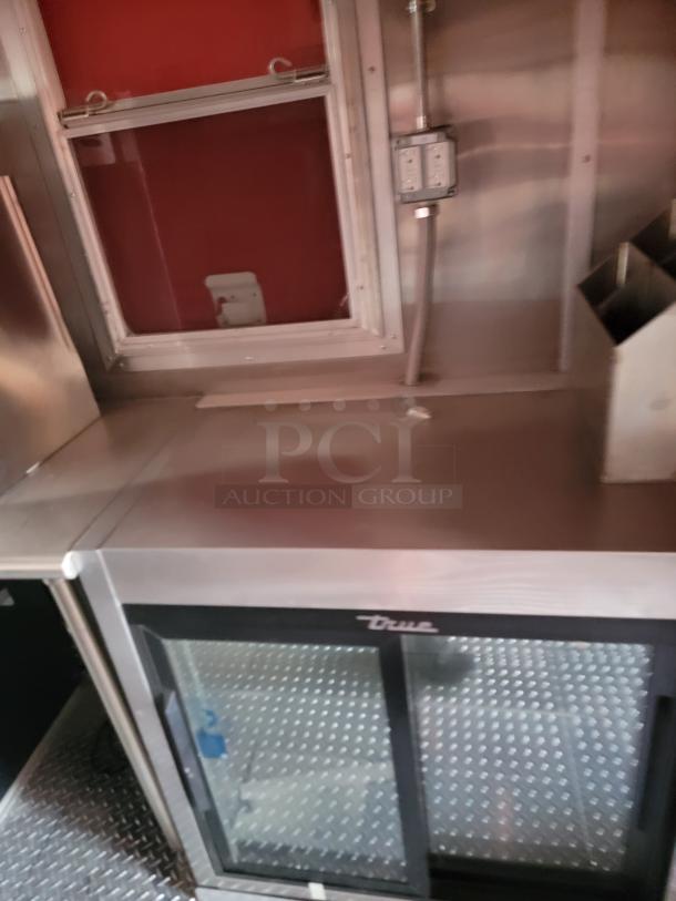 True commercial refrigerator with glass doors, stainless steel top, and red interior. Visible outlets and shelving unit. Good condition.