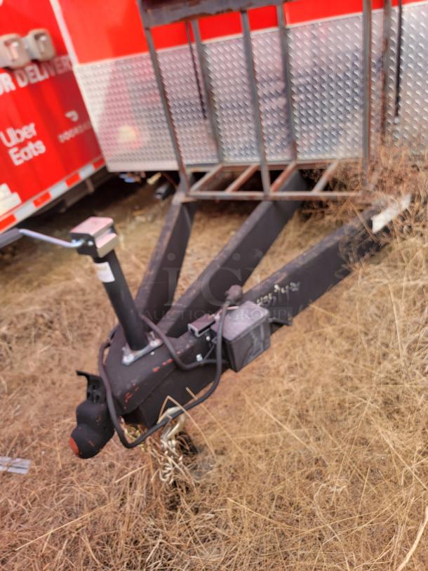 Trailer hitch with black metal frame and chains attached, resting in dry grass. Trailer in background with red and silver color scheme.