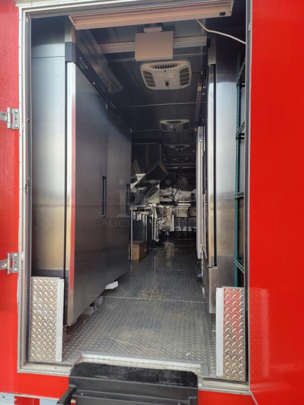 Interior view of a mobile kitchen trailer, featuring stainless steel appliances, ventilation system, and textured metal flooring.